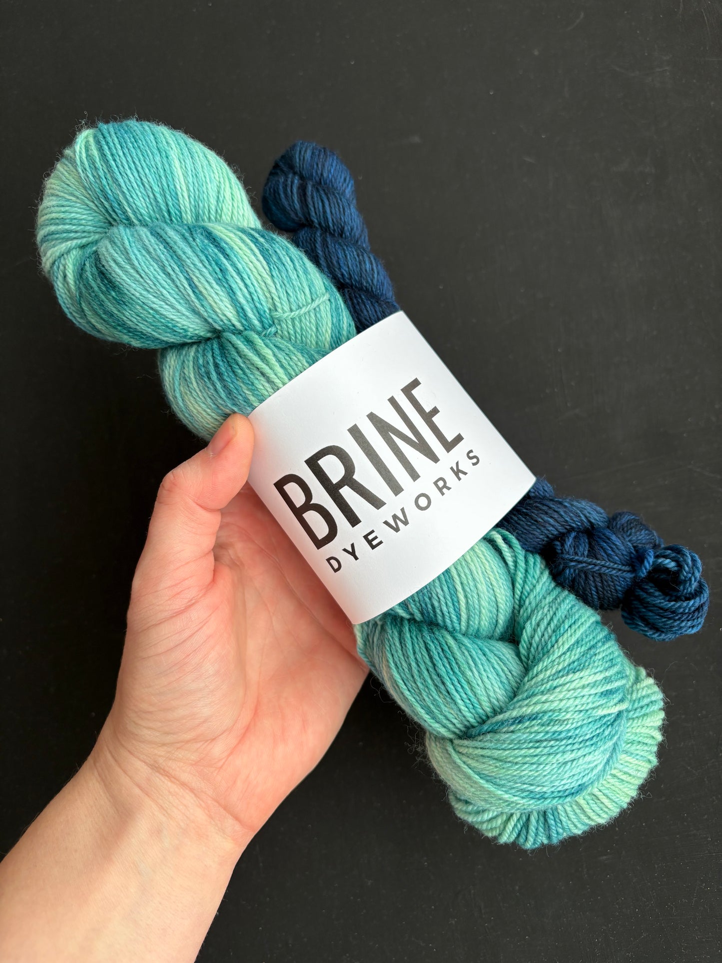 caucasian persoin's hand holding a sock set consisting of a green, teal and blue variegated skein of yarn with a mini skein of dark blue yarn with green undertones. Brand lable on yarn reads "Brine Dyeworks"