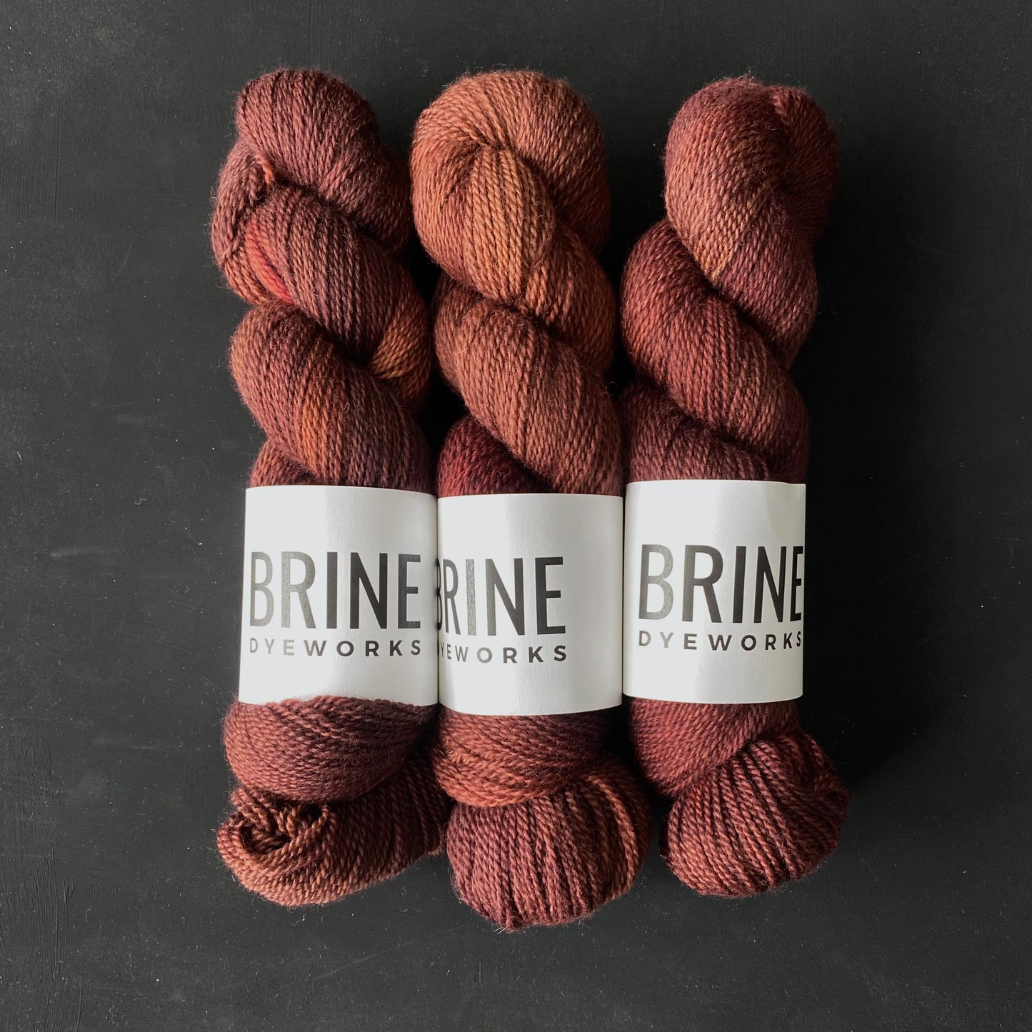 three skeins of reddish brown yarn with Brine Dyeworks labels on a dark background