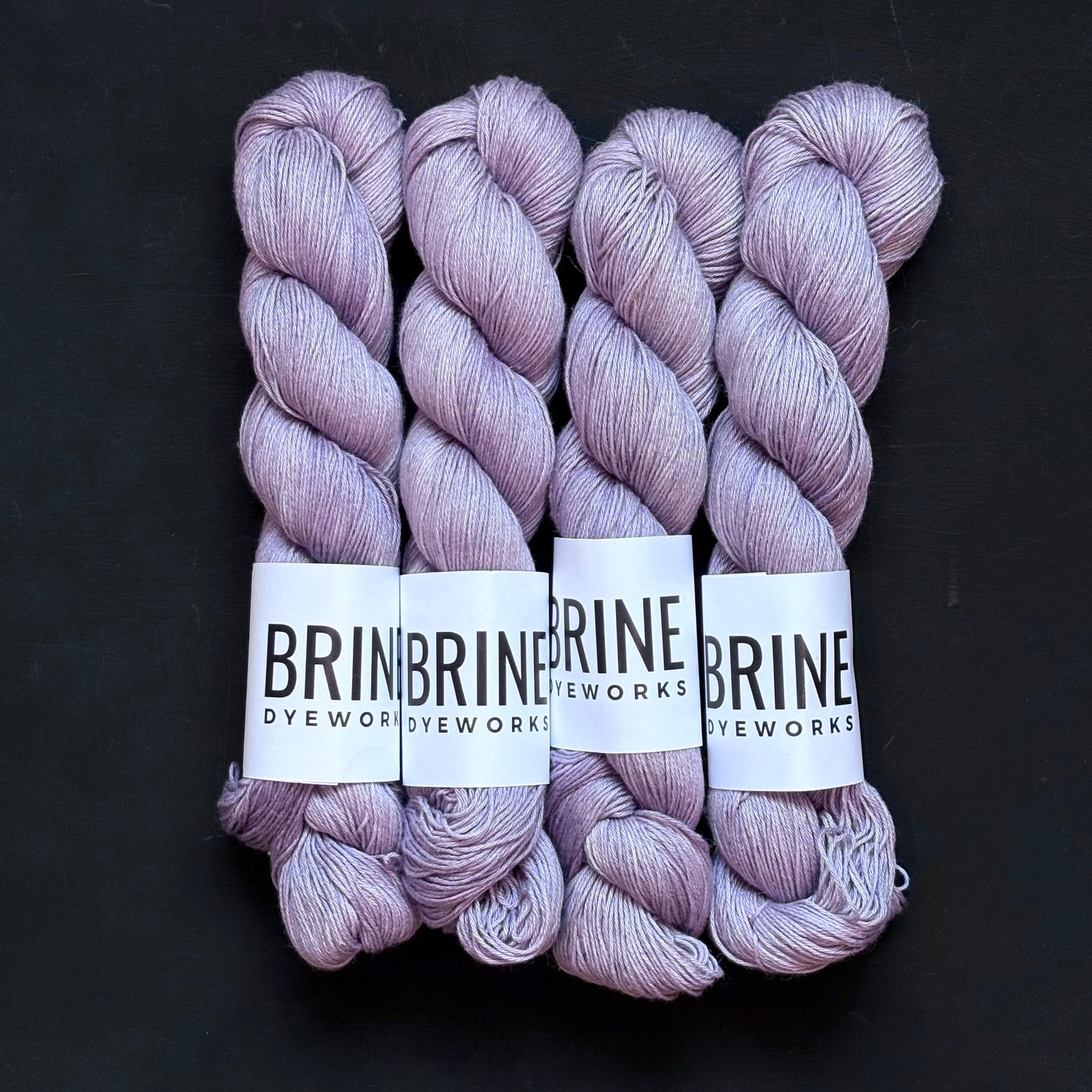 4 skeins of pale purple silk linen sport weight yarn with Brine Dyeworks labels on a dark back ground