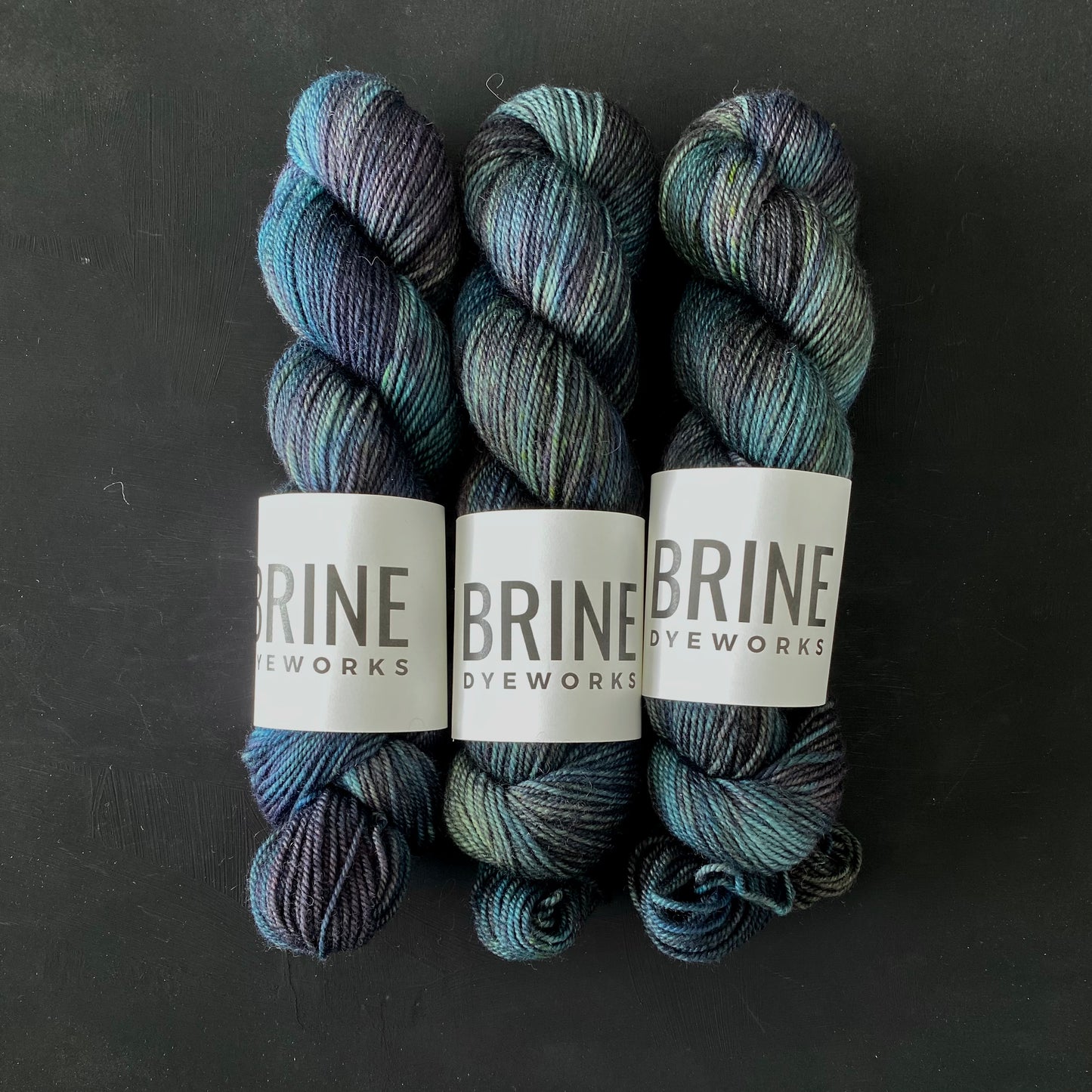 Three multicolored skeins of Brine Dyeworks yarn for knitting and other yarn crafts, on a dark background