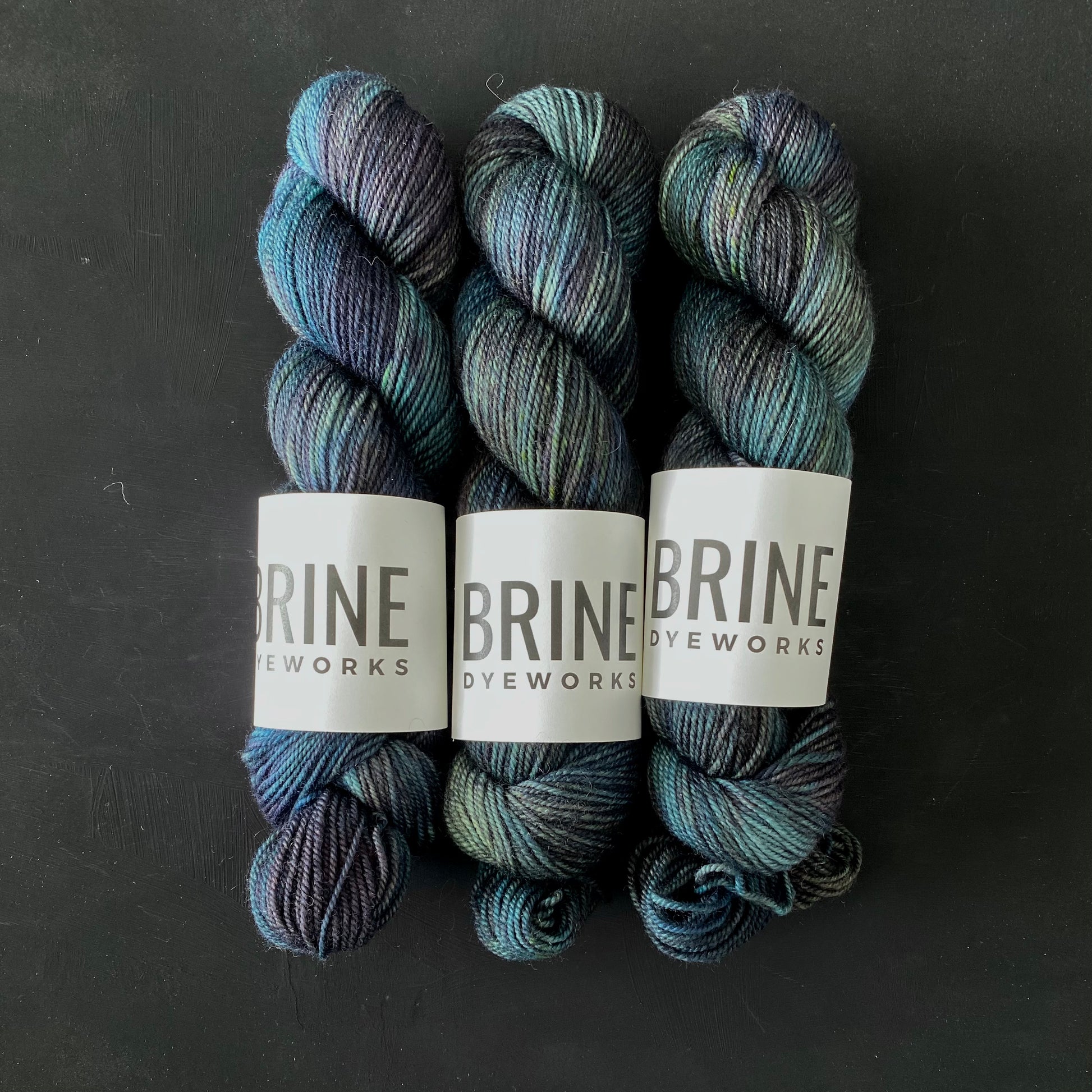 Three multicolored skeins of Brine Dyeworks yarn for knitting and other yarn crafts, on a dark background