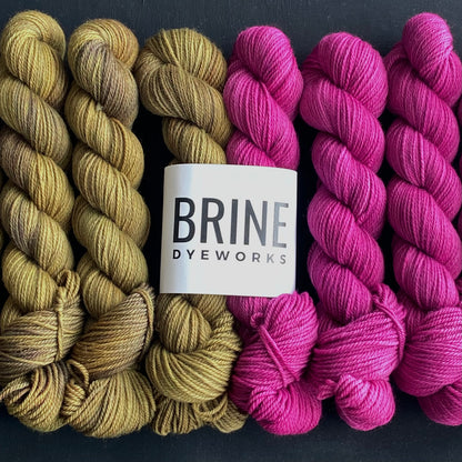 Yarn color combination 1: Pink Sapphire + Tarnish (orchid pink and dirty gold). Yarn is shown in 50g skeins and also packaged as a bundle with one of each color inside the Brine Dyeworks Logo packaging.