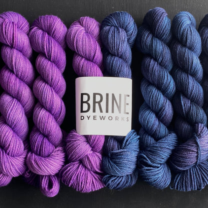 Yarn color combination 3: Deep + Thrills (dark blue and bright purple). Yarn is shown in 50g skeins and also packaged as a bundle with one of each color inside the Brine Dyeworks Logo packaging.