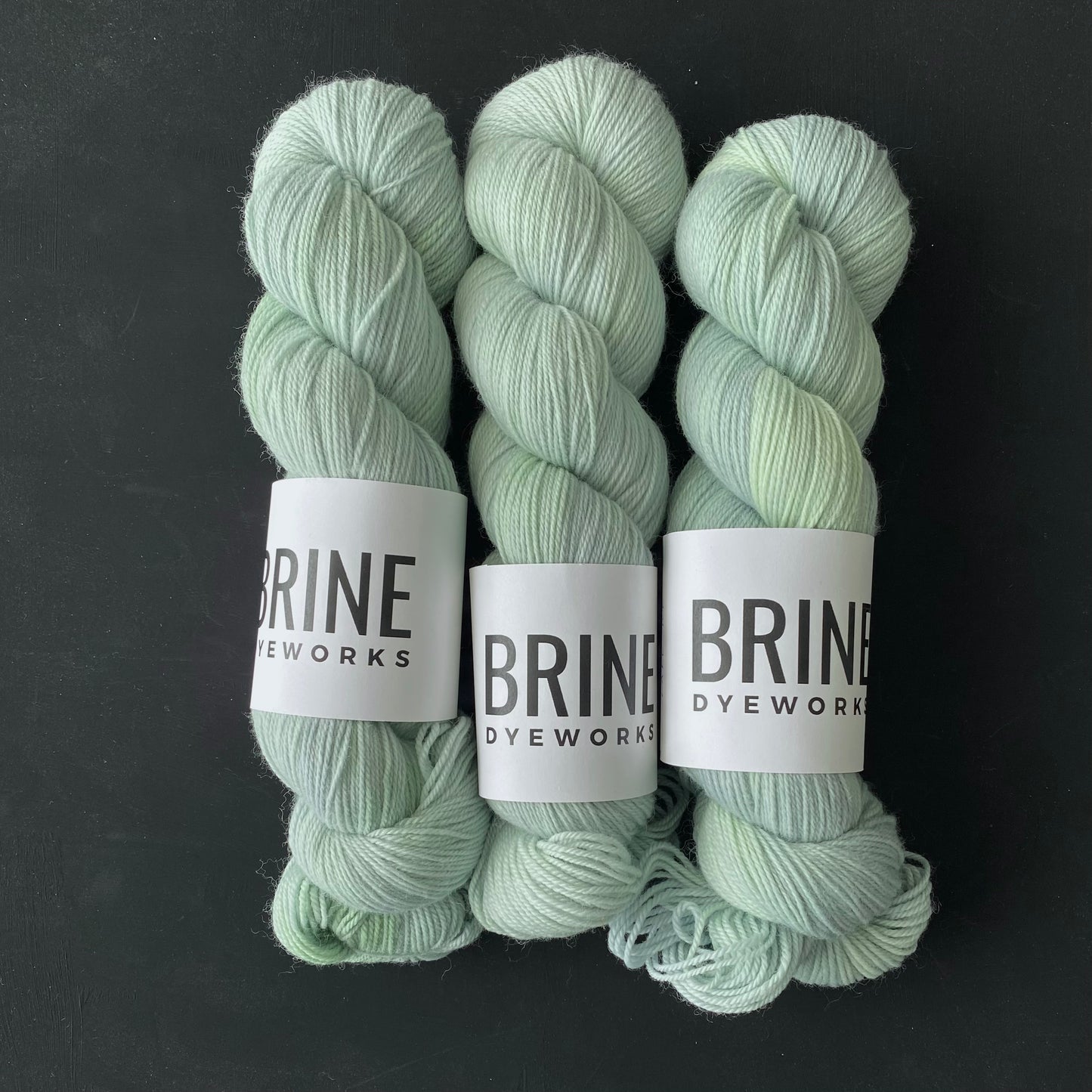 Three skeins of light green yarn arranged vertically on a dark background, with labels indicating the brand 'BRINE Dyeworks'.