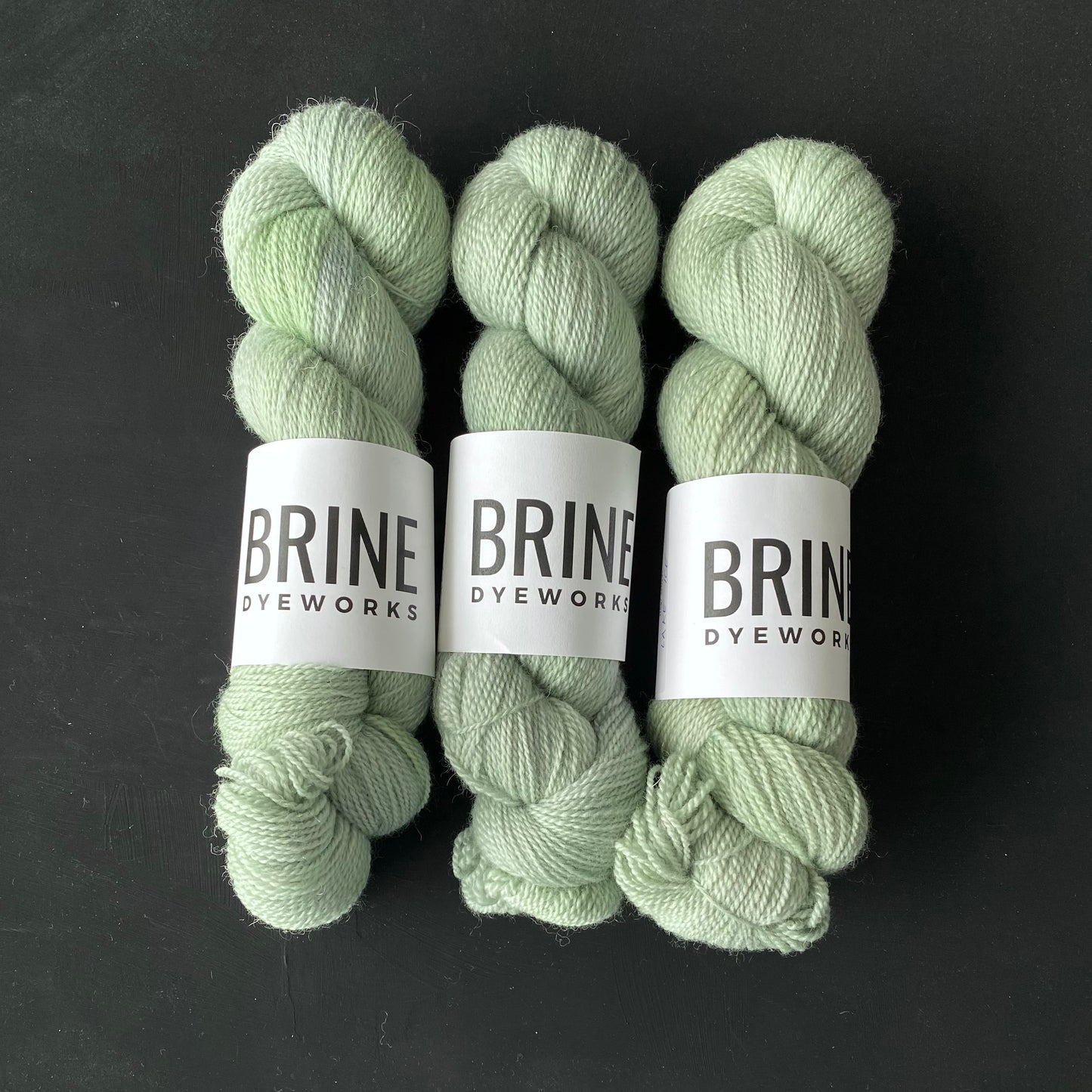 Three skeins of untreated blue faced leicester wool yarn in a light green color, labeled with Brine Dyeworks branding.
