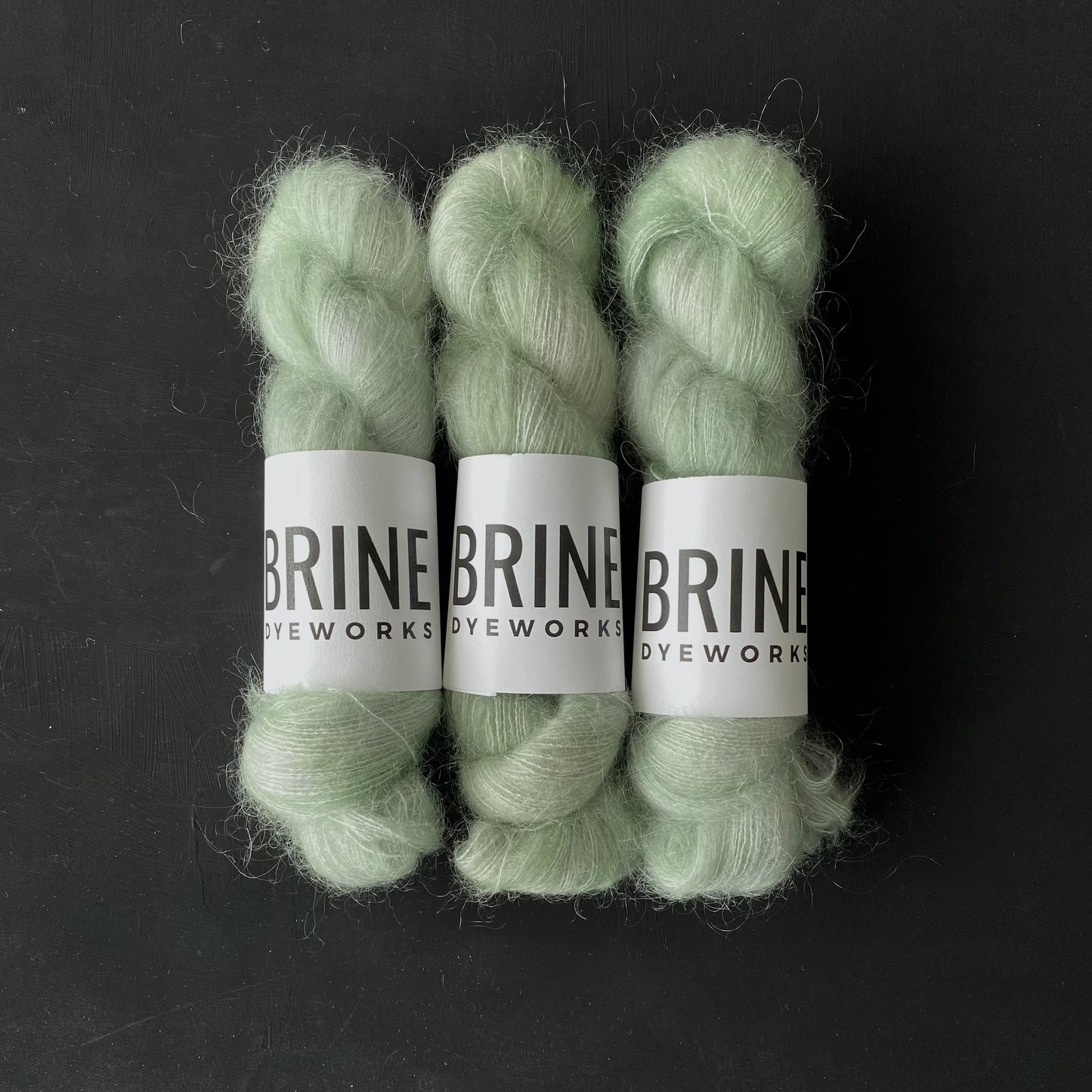 Three Skeins of mohair silk lace yarn in a light green color with Brine Dyeworks labels and a dark background