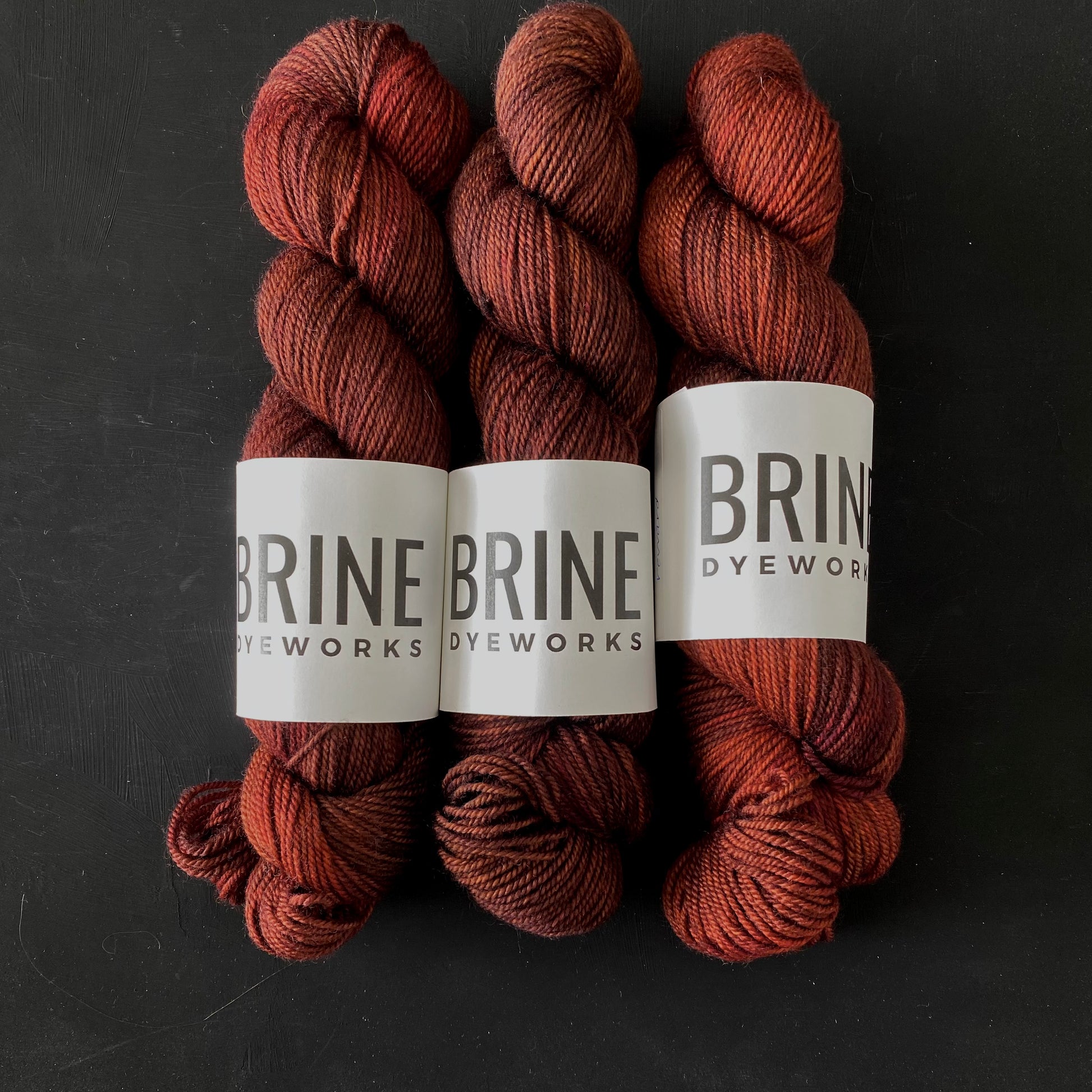 Three skeins of reddish brown yarn for knitting with Brine Dyeworks labels on a dark background
