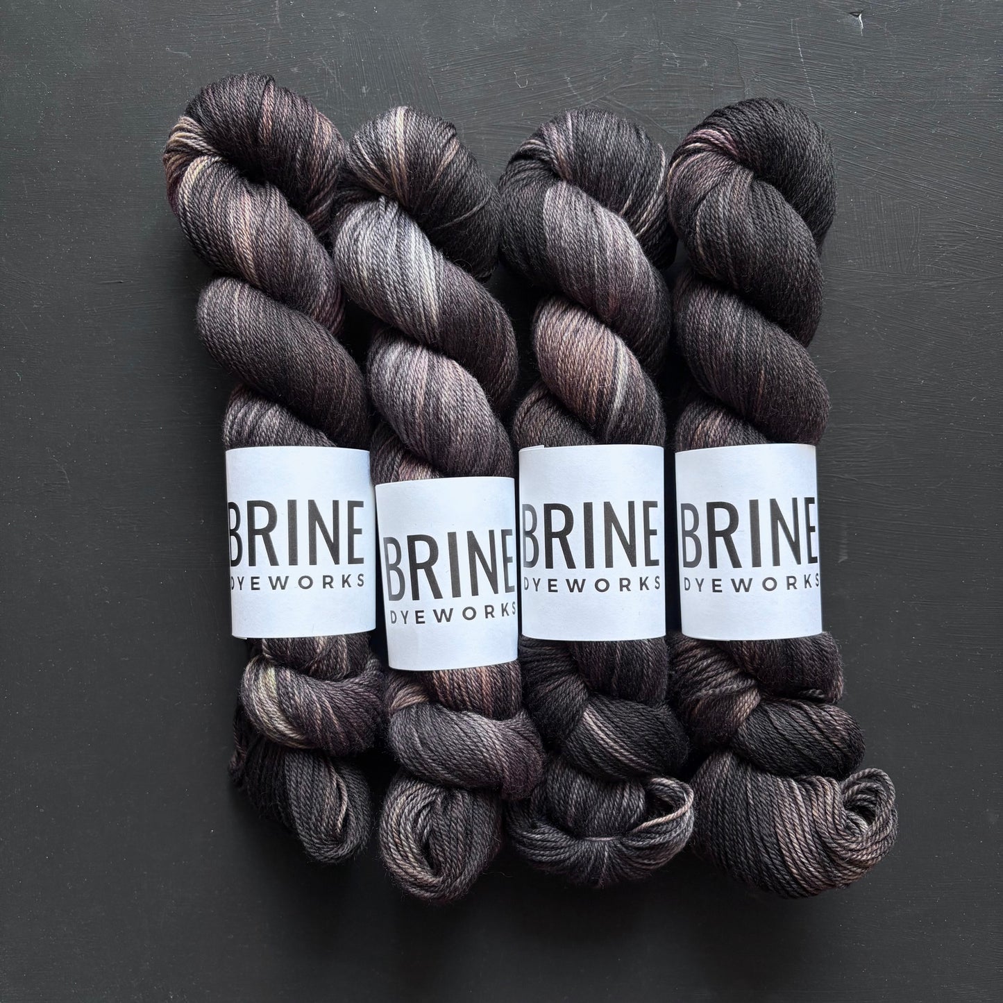 Four skeins of dark brown yarn with varying shades of black, brown and grey, wrapped in BRINE DYEWORKS labels on a dark background